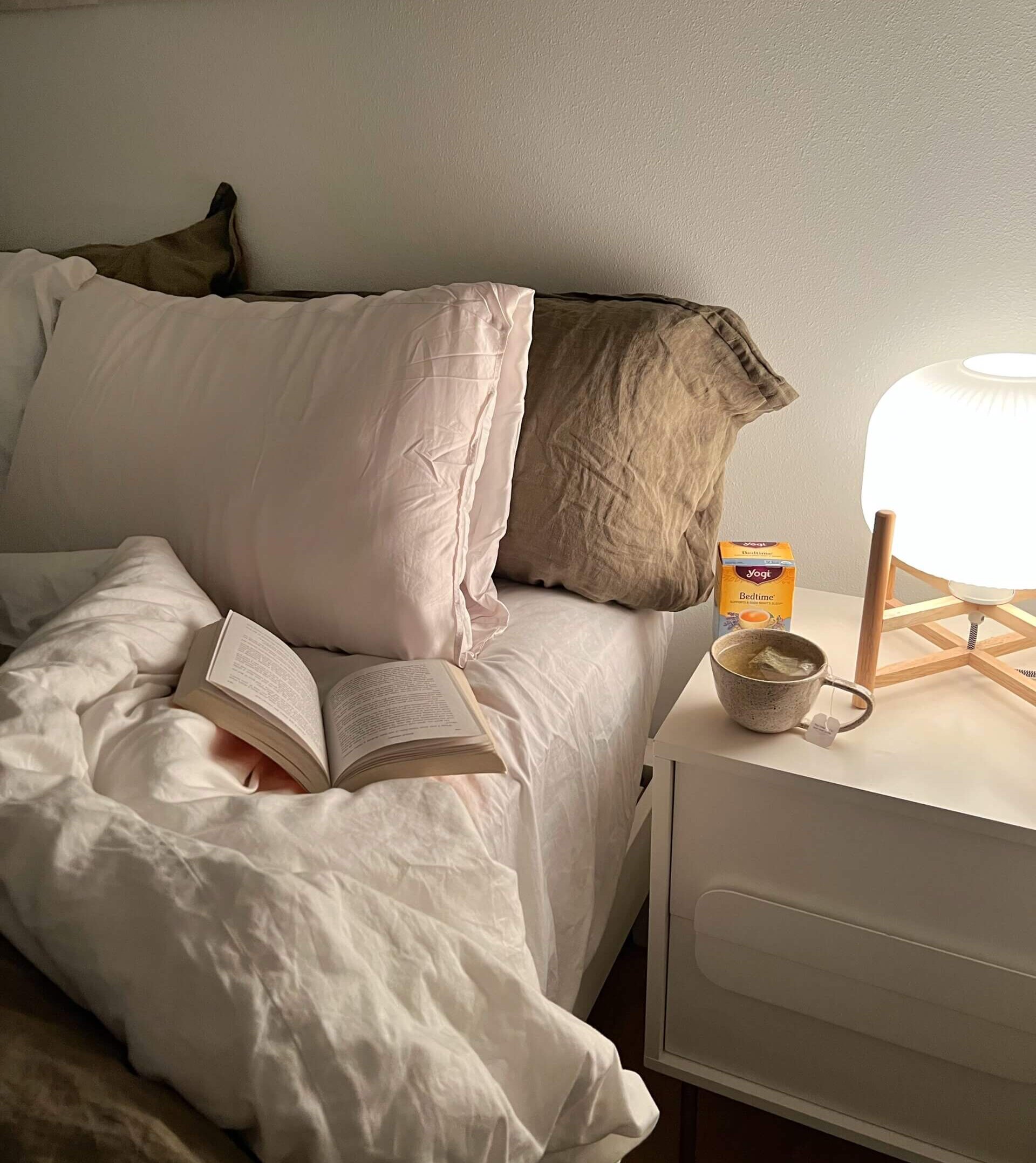Bed with open book resting on it. A cup of tea sits on the nightstand