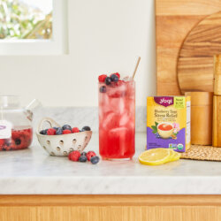 A glass of Mixed Berry Iced Tea Cooler sits on a counter next to a box of Yogi Blueberry Sage Stress Relief tea and a bowl of fresh berries.