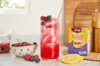 a glass of Mixed Berry Iced Tea Cooler sits on a counter next to a box of Yogi Blueberry Sage Stress Relief tea, a bowl of fresh blueberries and raspberries, and sliced of lemon.