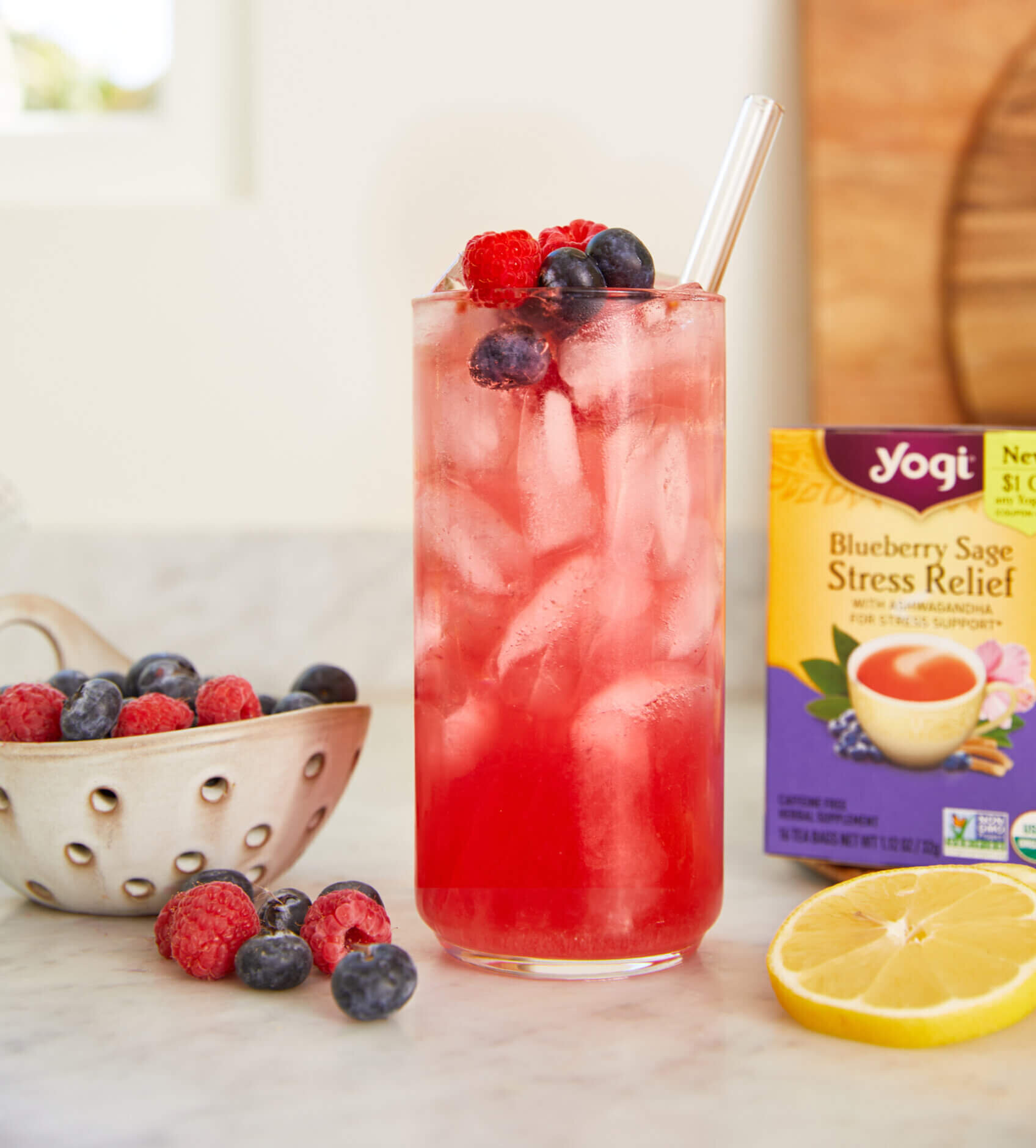a glass of Mixed Berry Iced Tea Cooler sits on a counter next to a box of Yogi Blueberry Sage Stress Relief tea, a bowl of fresh blueberries and raspberries, and sliced of lemon.