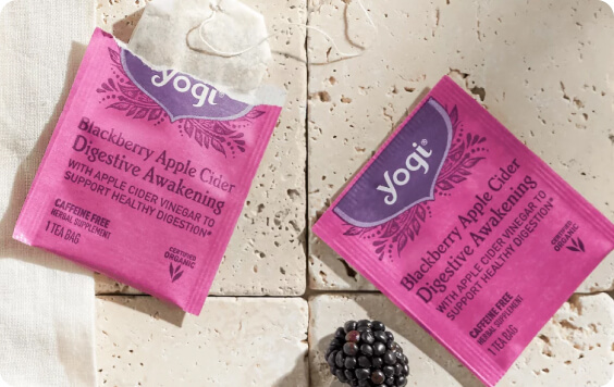 Two Yogi tea bags of Blackberry Apple Cider Digestive Awakening on a textured surface, one torn open, with a fresh blackberry.