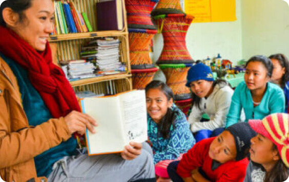 A woman reads to children in a colorful classroom, with engaged kids wearing knit hats, suggesting a storytelling session.